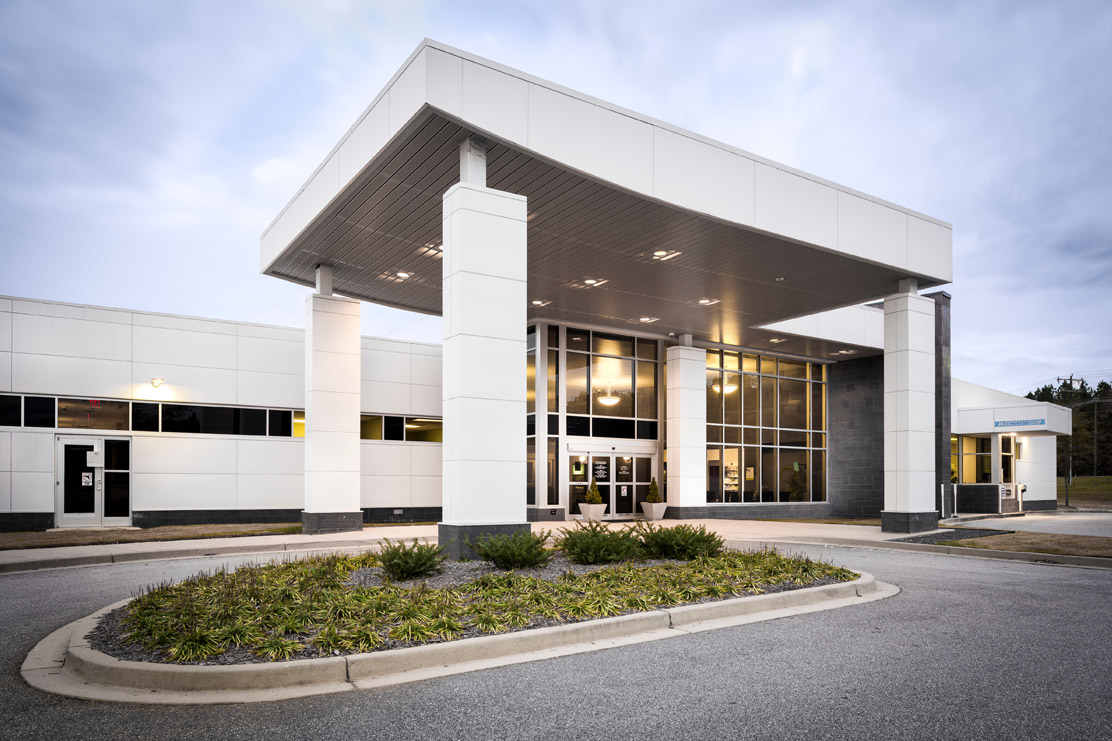 The Associate Family Health Center is a white building with a large windowed covered entrance. The pharmacy drive-up window is on the right side of the building. 