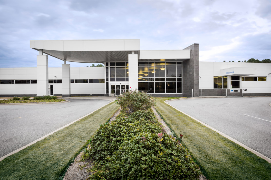 The Associate Family Health Center is a white building with a large windowed covered entrance. The pharmacy drive-up window is on the right side of the building. 