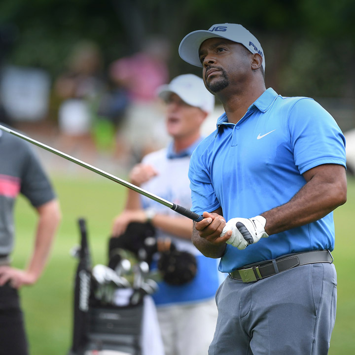 Golfer in a blue shirt watches as his golf ball prepares to land. His golf caddy in the background attentively watches. 