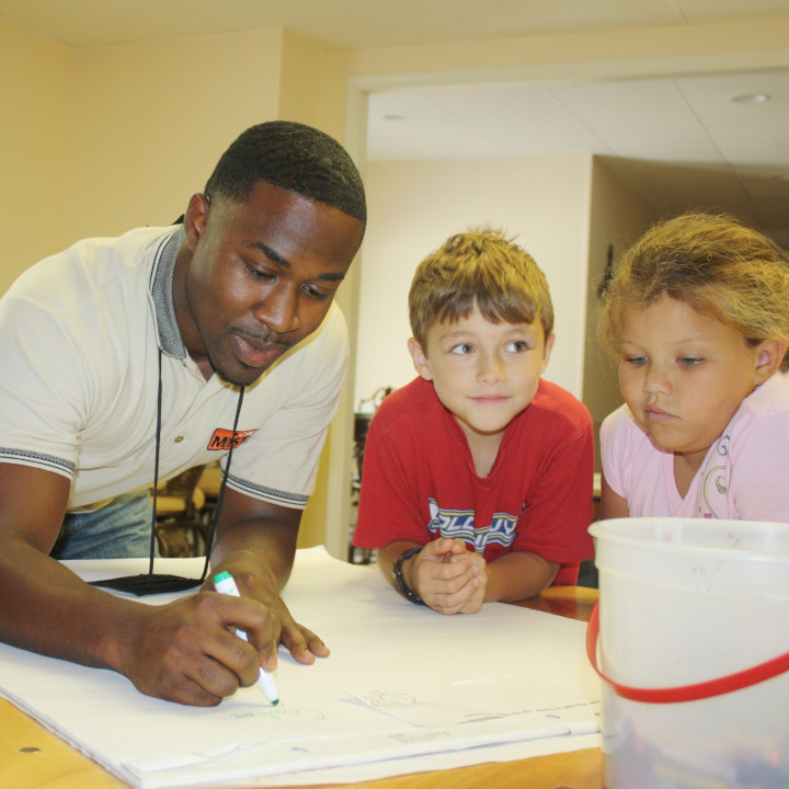 A teacher  in the Call Me Mister program illustrates a lesson to two attentive children in a classroom. 