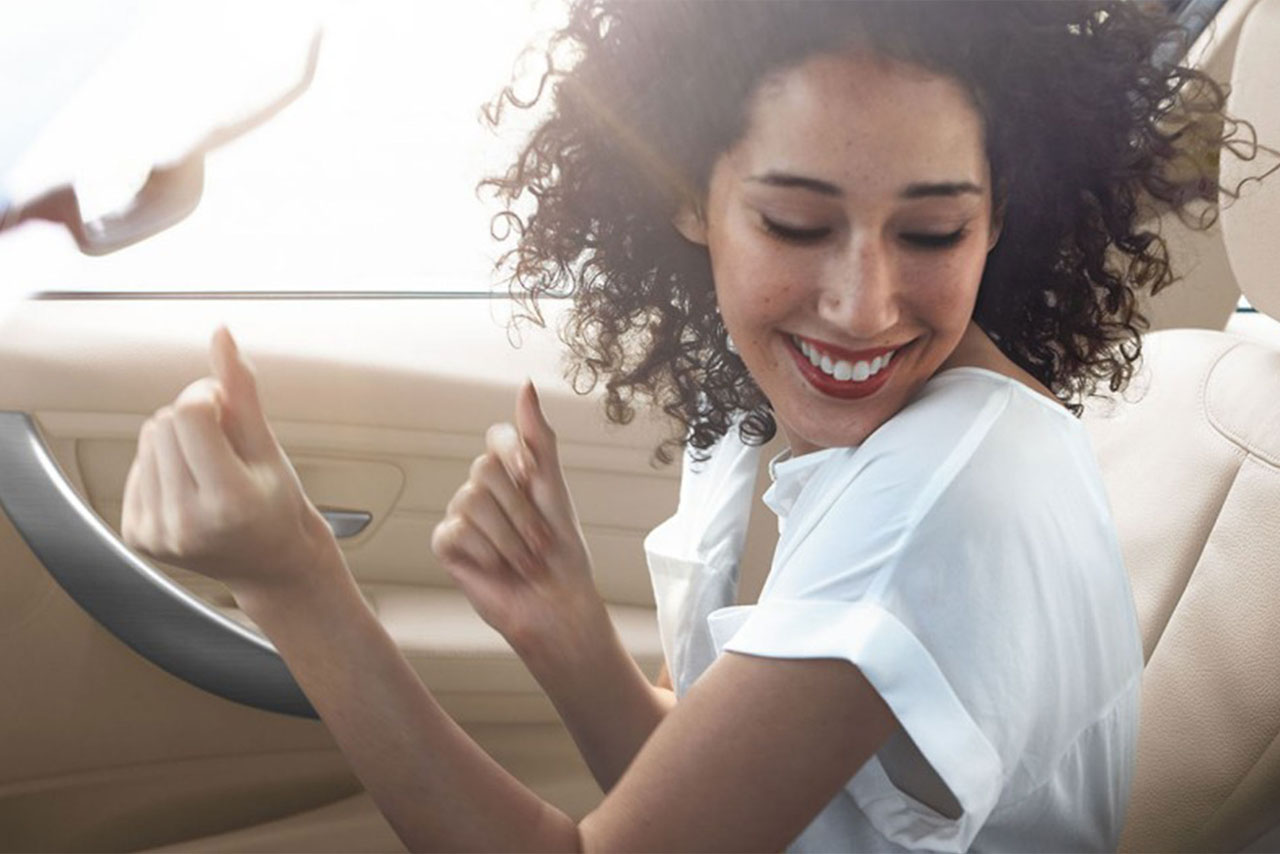 A woman smiles happily while sitting in the driver's seat of a car, reflecting a sense of enjoyment and readiness.